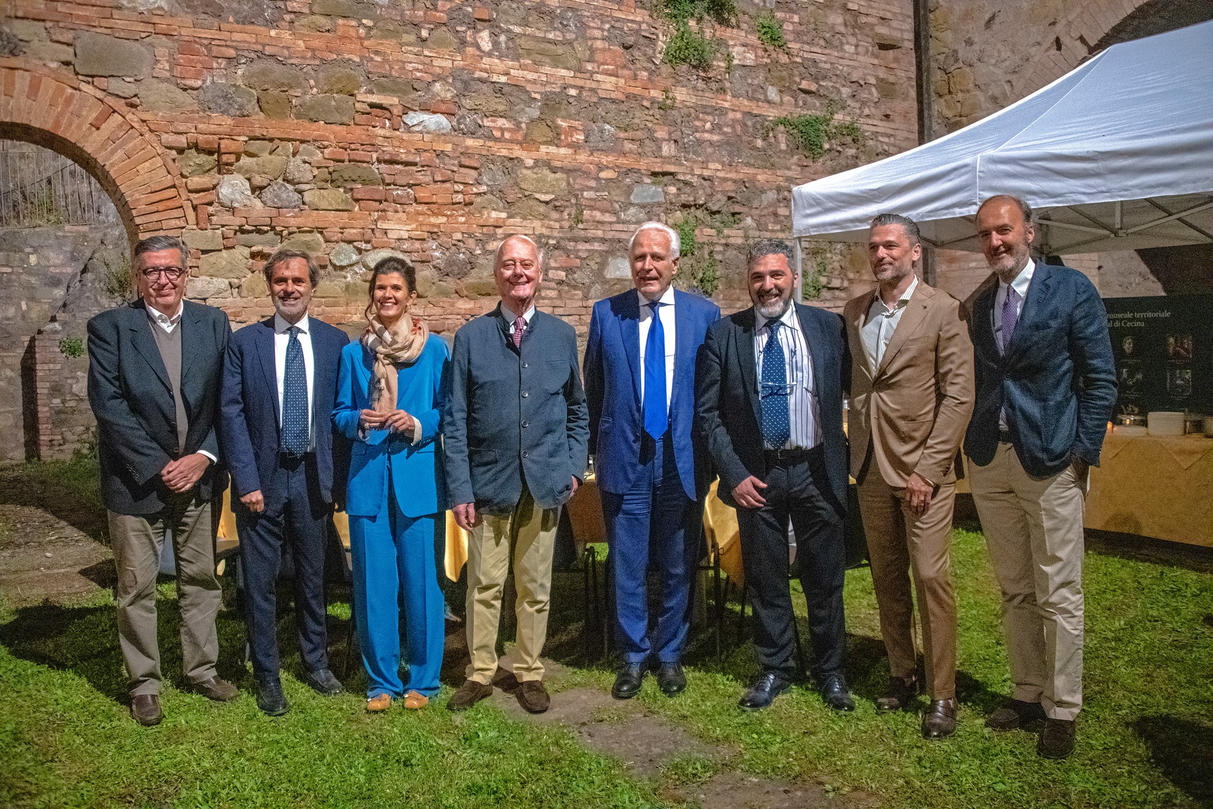 Group of people in formal attire standing outdoors near a stone wall and a white canopy