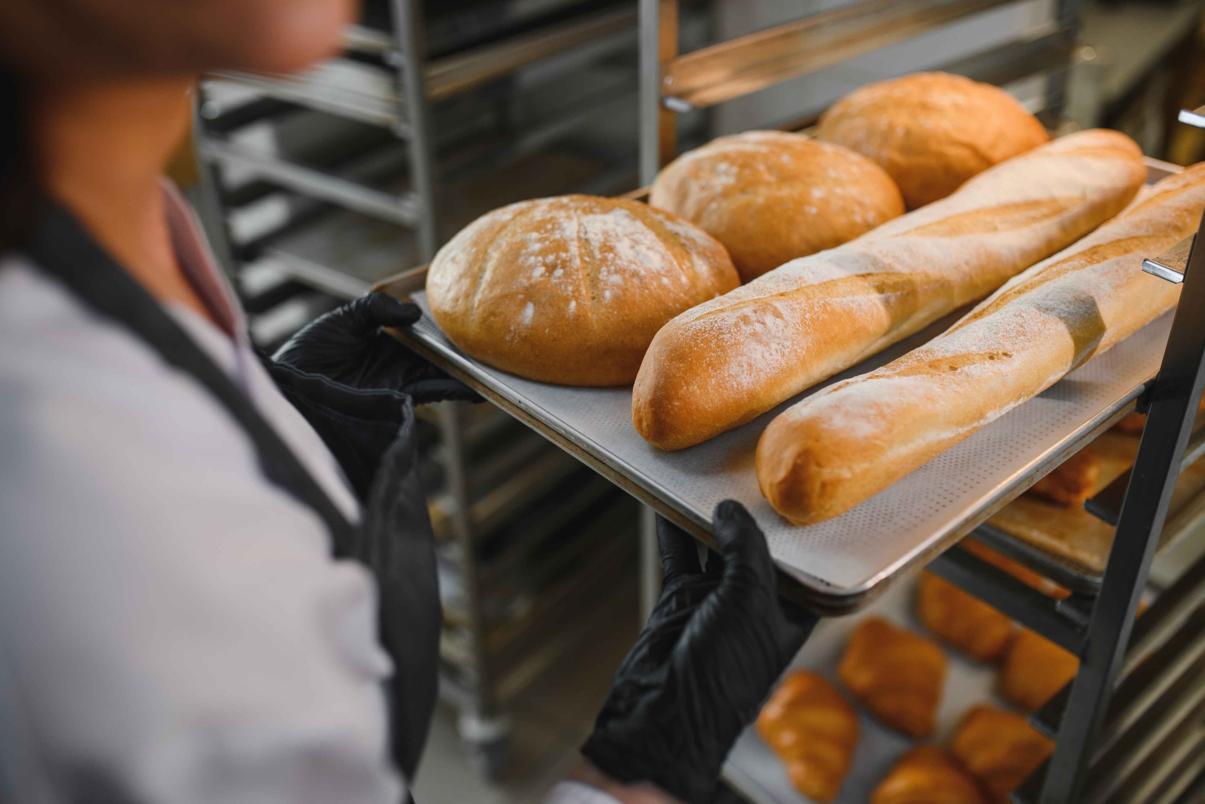 Fornaio che indossa guanti neri che tengono il vassoio di metallo con le pagnotte appena cotte e le baguette nella cucina del panificio