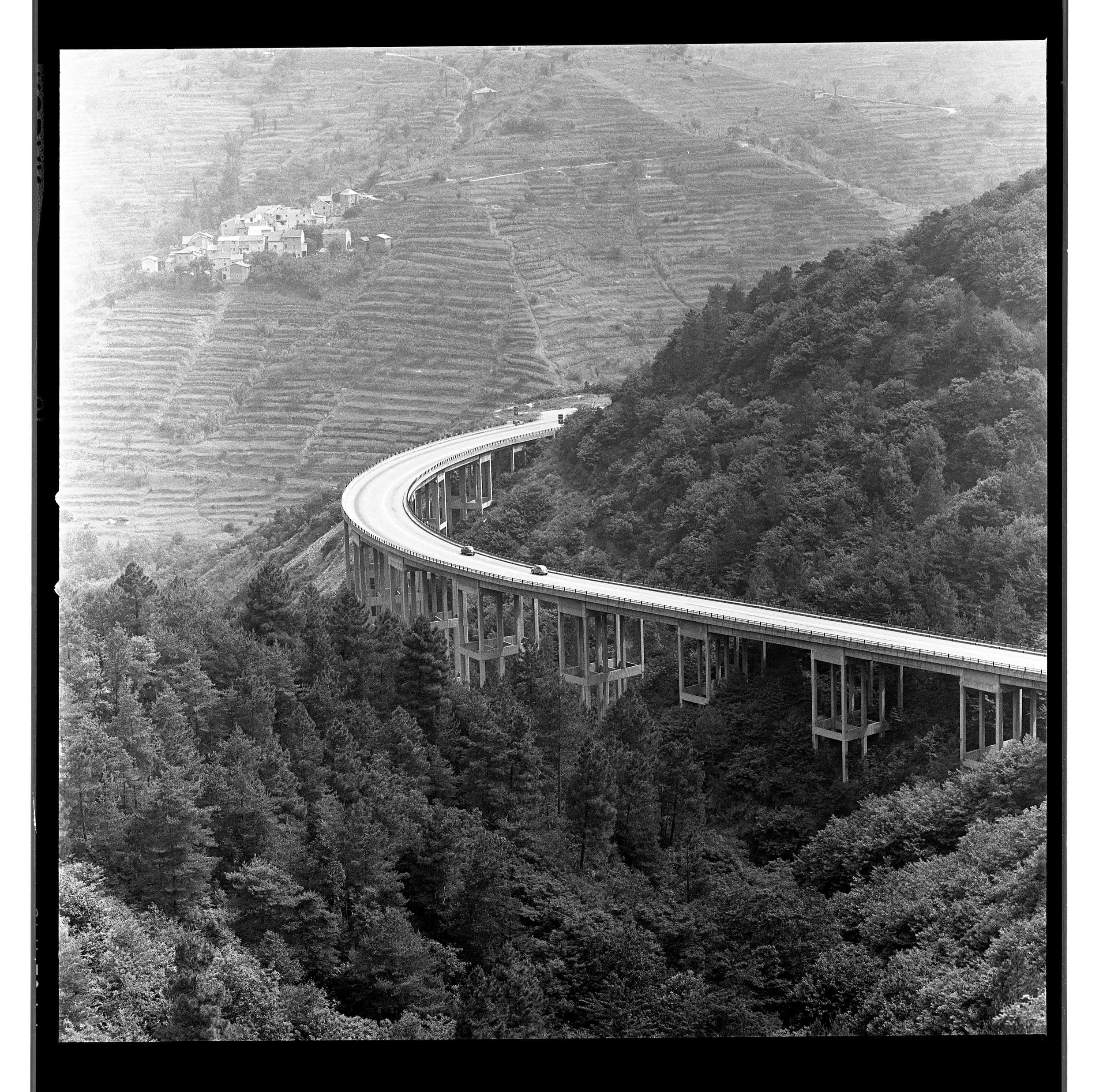 Black-and-white photo of a curving elevated highway cutting through forested hills and terraced slopes