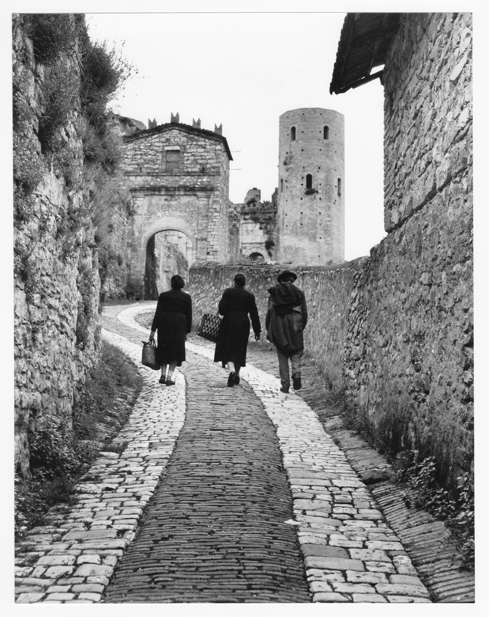 Black-and-white photo of three people walking along a cobblestone path toward a medieval stone gate and tower