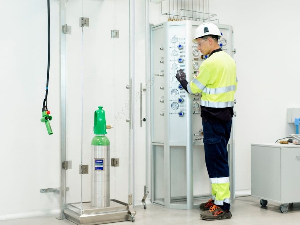 Industrial technician in high-visibility gear operating gas control panel near cylinder in safety enclosure